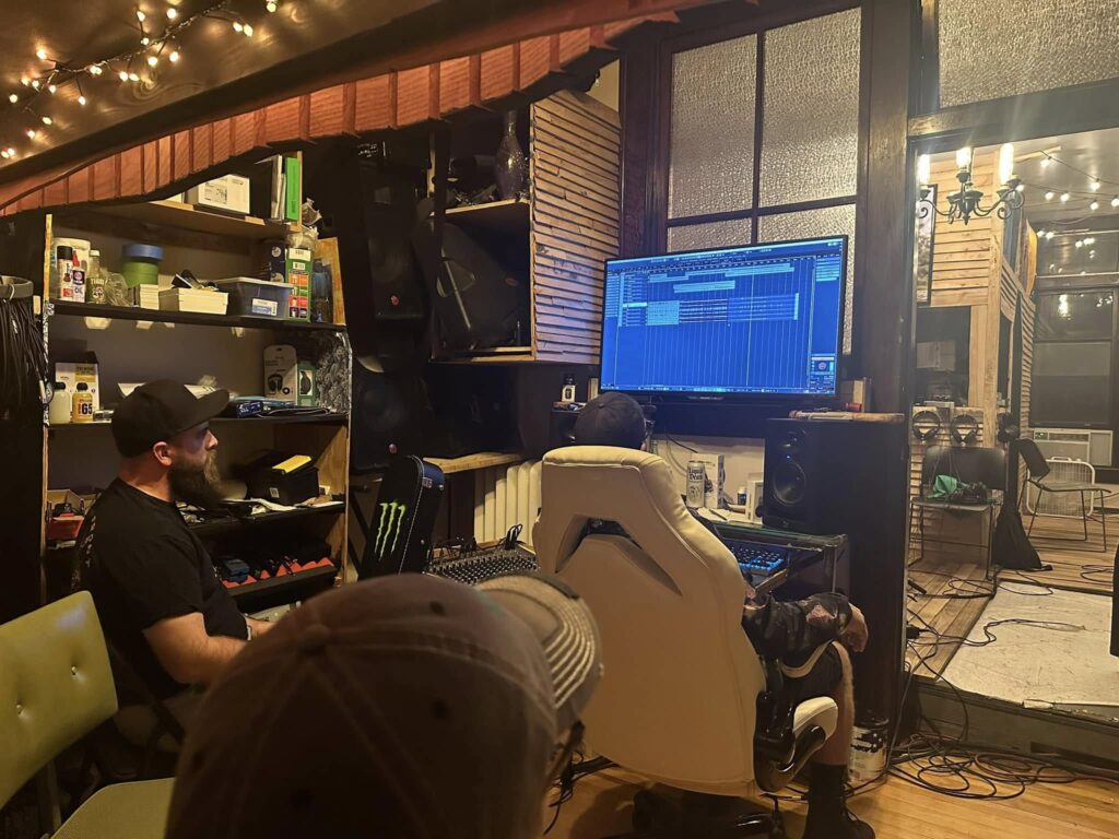 Two people work at a music studio desk with a large monitor displaying audio production software and equipment.