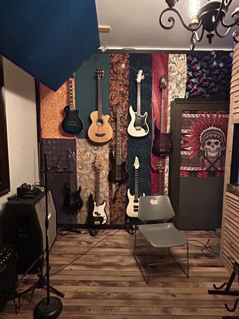 Six guitars and basses hang on a decorated wall in a music studio with a chair and amplifiers on a wooden floor.