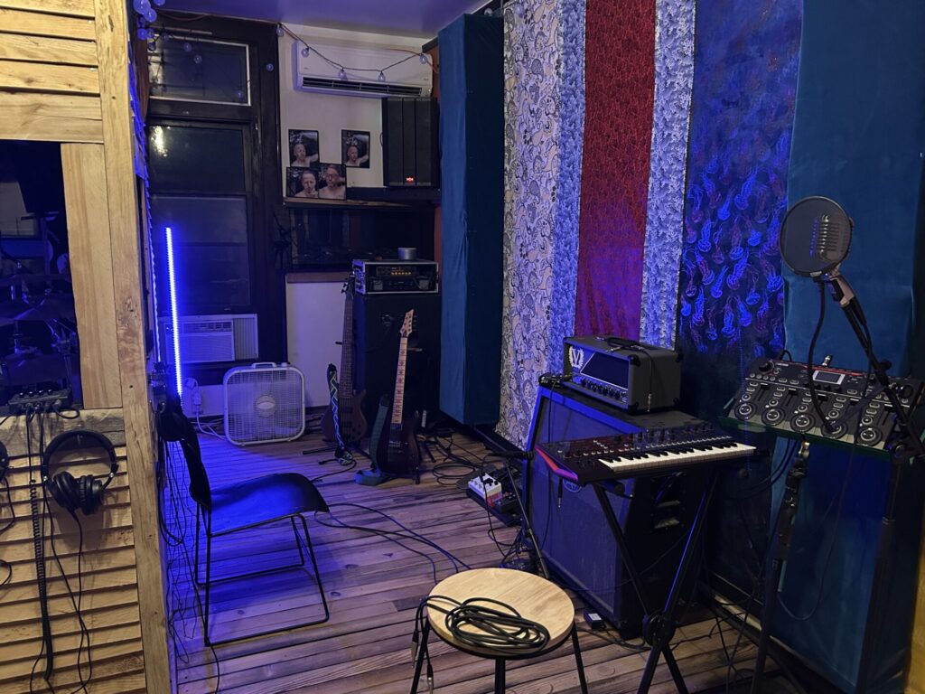 A recording studio room with musical instruments, microphone, keyboard, amplifier, headphones, and soundproofing panels.